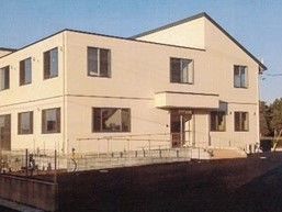 2階建ての建物はベージュを基調としている。周辺には緑が多く、右側には山もある。