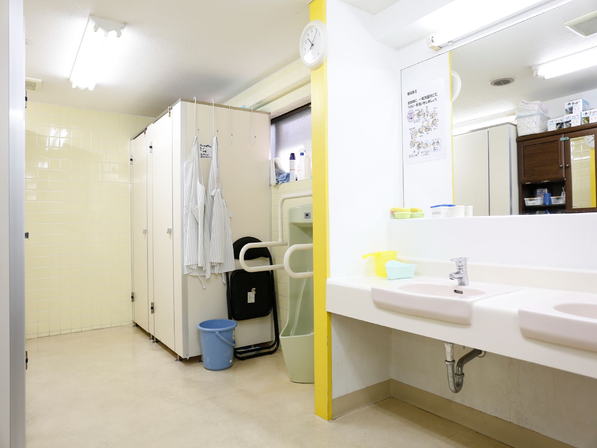 個室と、男性用小便器と洗面台が2つ見える。洗面台の上には大きな鏡があり柱には時計が掛かっている。男性用小便器の両側には手すりが付いている。