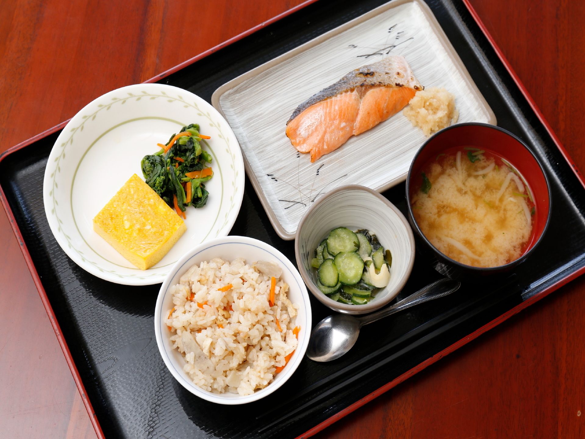テーブルの上には食事の載ったお盆が置かれている。写真には色とりどりの卵焼きや焼き鮭、酢の物や混ぜご飯、味噌汁がある。