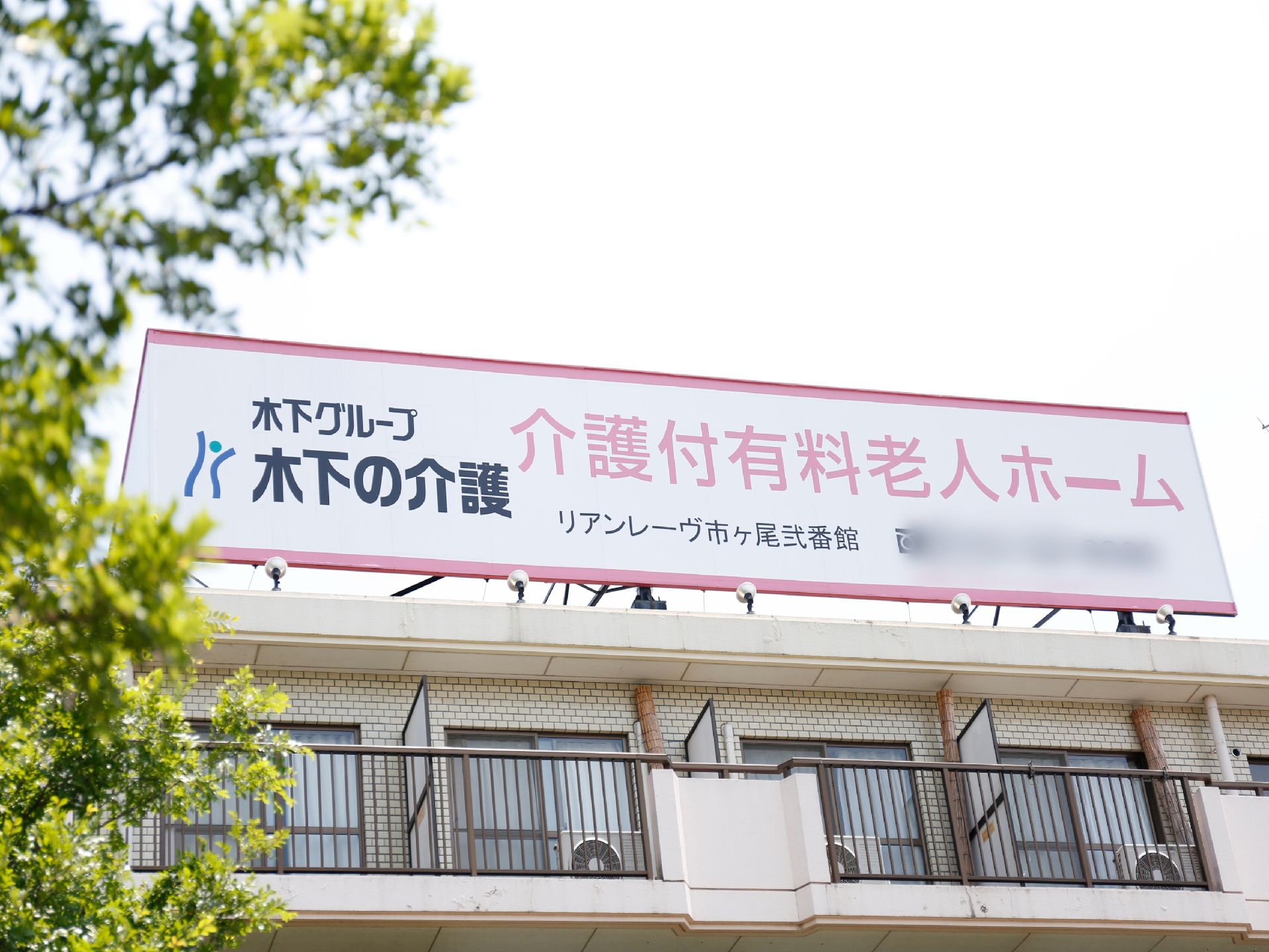 施設の建物の上に、ピンク色と白色の大きい看板が建っており、施設名や施設形態、電話番号や会社名が書かれている。木々が左の方に映っている。