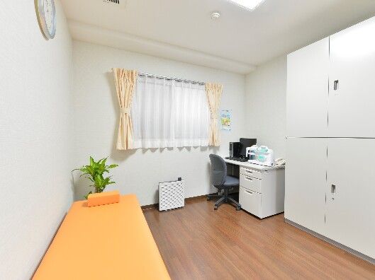 布団のないオレンジのベッドやデスクが設置されたフローリングの部屋。レースカーテンとカーテンが付いた窓がある。