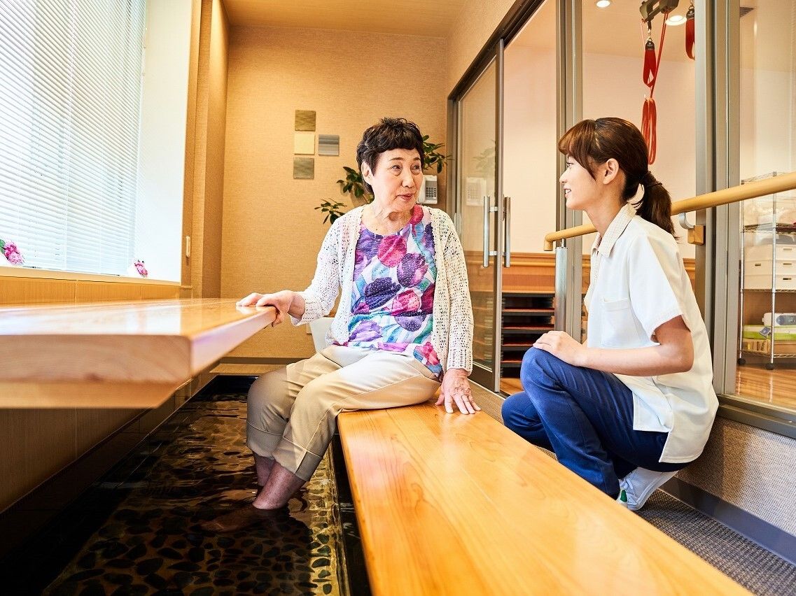 車いすにのる男性とそれを押している女性スタッフ。両側に自然が広がる遊歩道の写真
