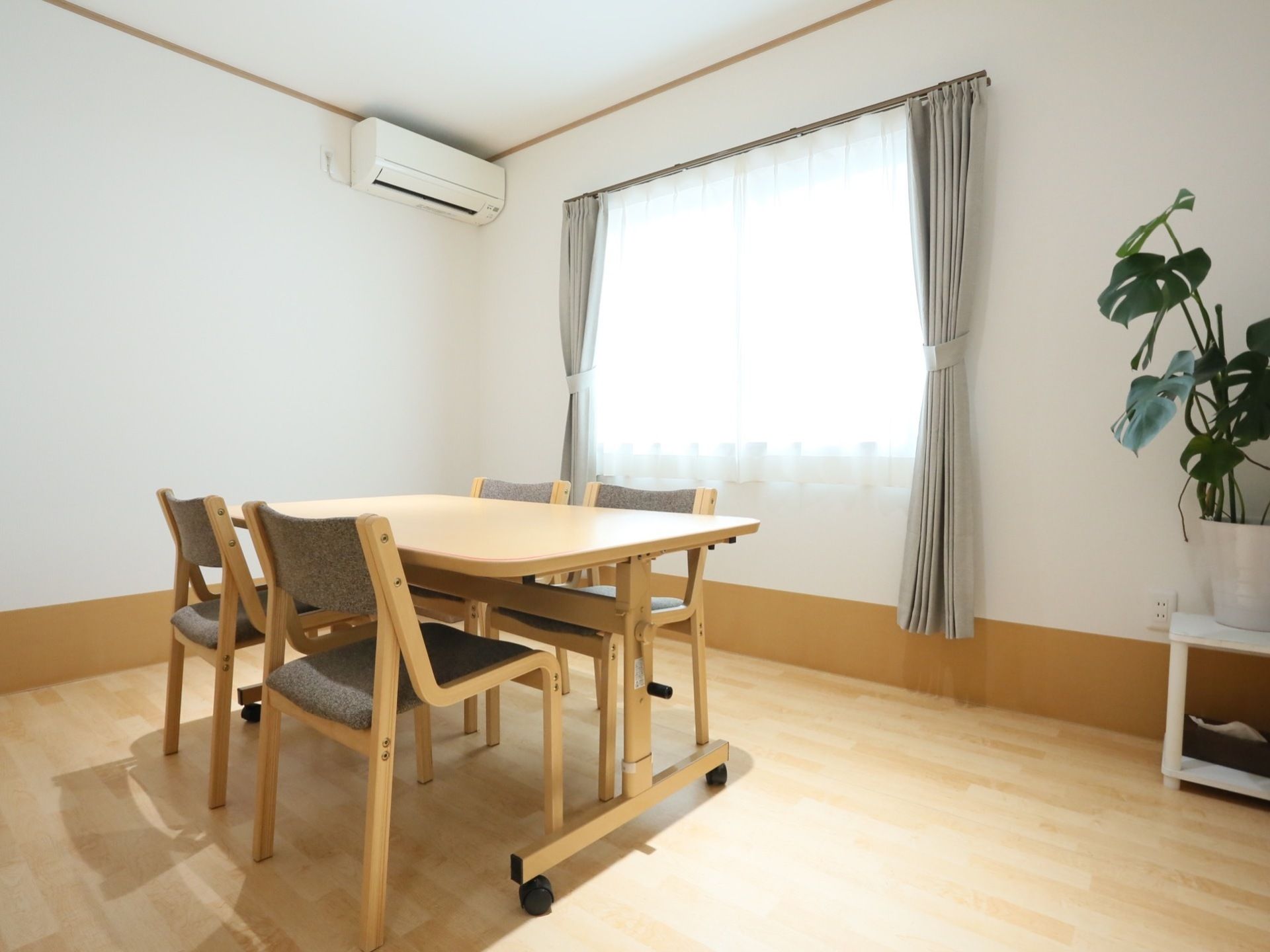 部屋の中央に４人がけテーブルが用意してある。窓があり、室内にはエアコンが取りつけてある。白い台に観葉植物を飾っている。