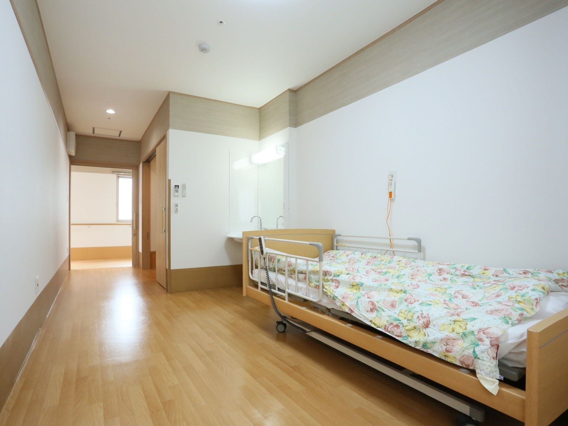 白い壁と木目調のフローリングのシンプルで明るい長方形の居室である。壁には緊急呼び出しボタンが設置されている。