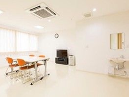 壁際にはテレビが置かれ、中央に4人掛けのテーブルセットがあり、車いす対応の洗面所が設置されている。