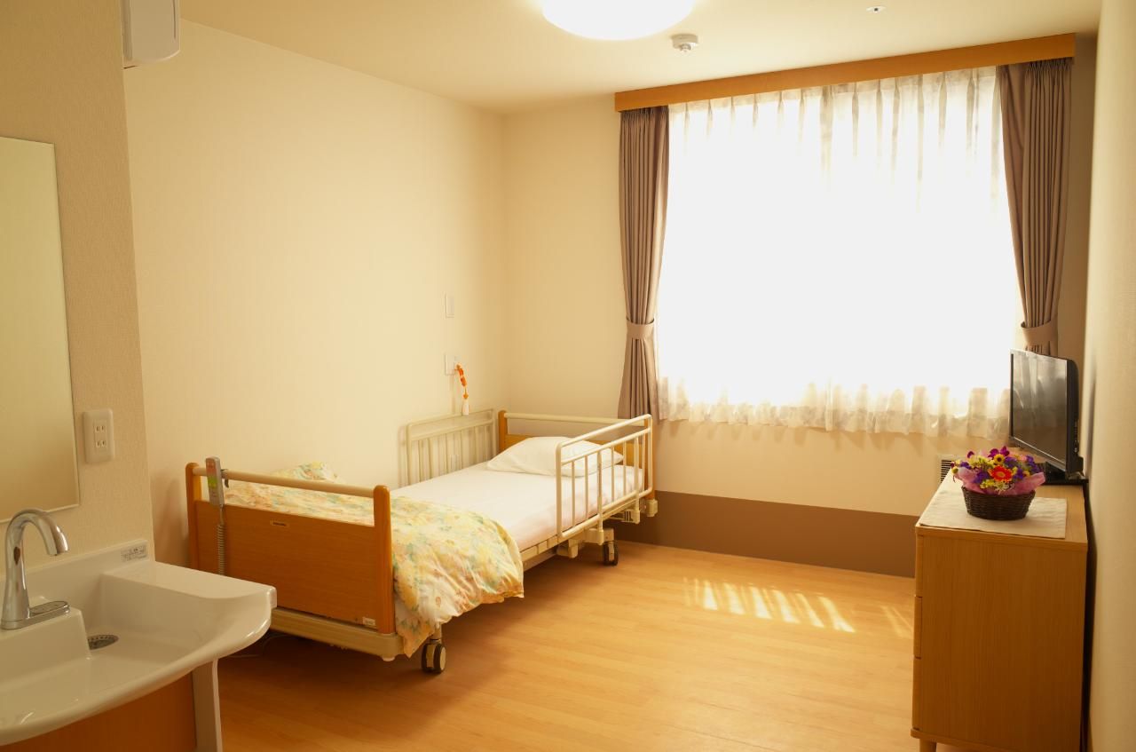 原っぱのような場所をブルーのエプロンを付けた若い女性スタッフに付き添われて歩いている高齢女性。
