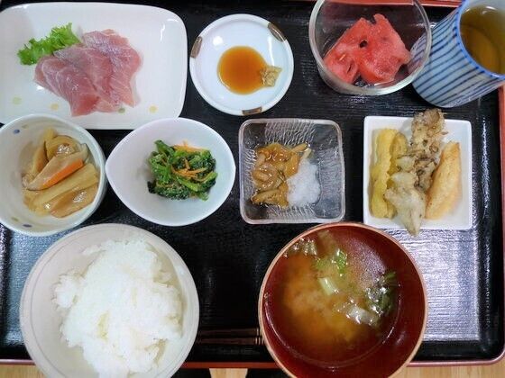 刺身になめこの大根おろし掛け、数種類の異なる野菜の煮ものに、白いご飯に味噌汁、そしてスイカとお茶が黒いトレーの上に並んでいる。
