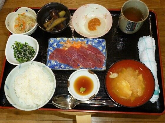 黒いトレーの上には白いご飯に味噌汁、刺身に野菜の和え物、風呂吹き大根、なますなどが所狭しと並べられ、おしぼりが添えられている。