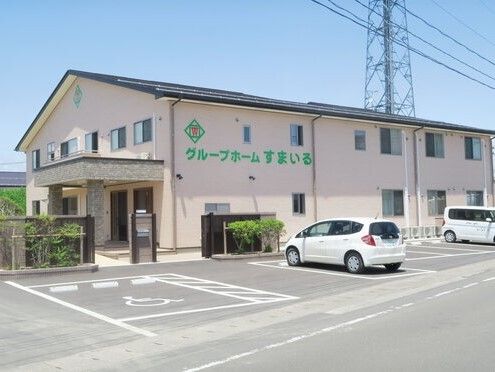 大きな駐車場には白い車が2台止まっていて、2階建ての建物の外壁には緑の文字でホーム名が書かれ、玄関には屋根が付いている。日当たりがいい場所にある。