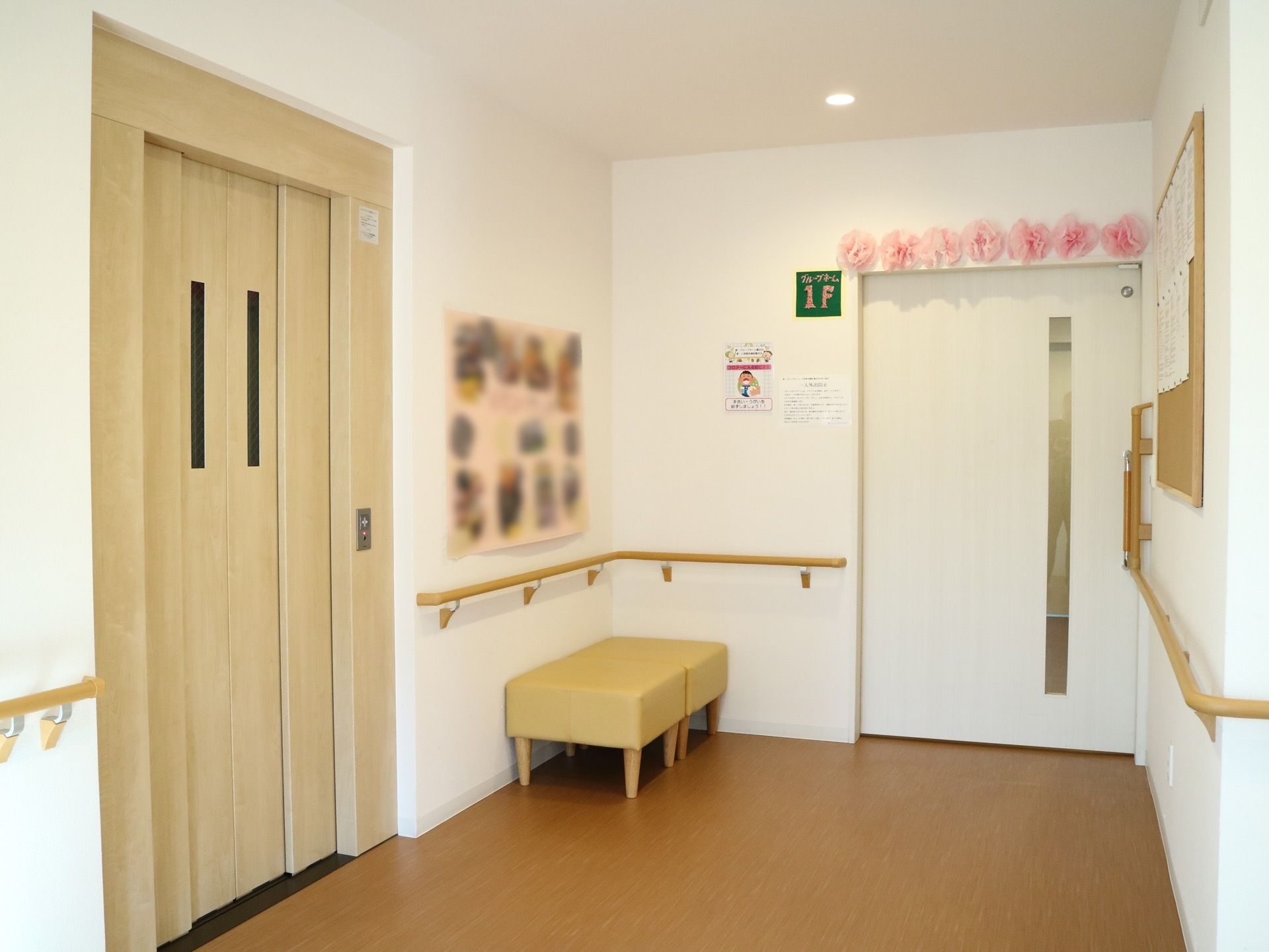エレベーターがある。乗降口の近くに腰かけが２つ並べて置かれている。その後ろの壁に写真が貼られている。
