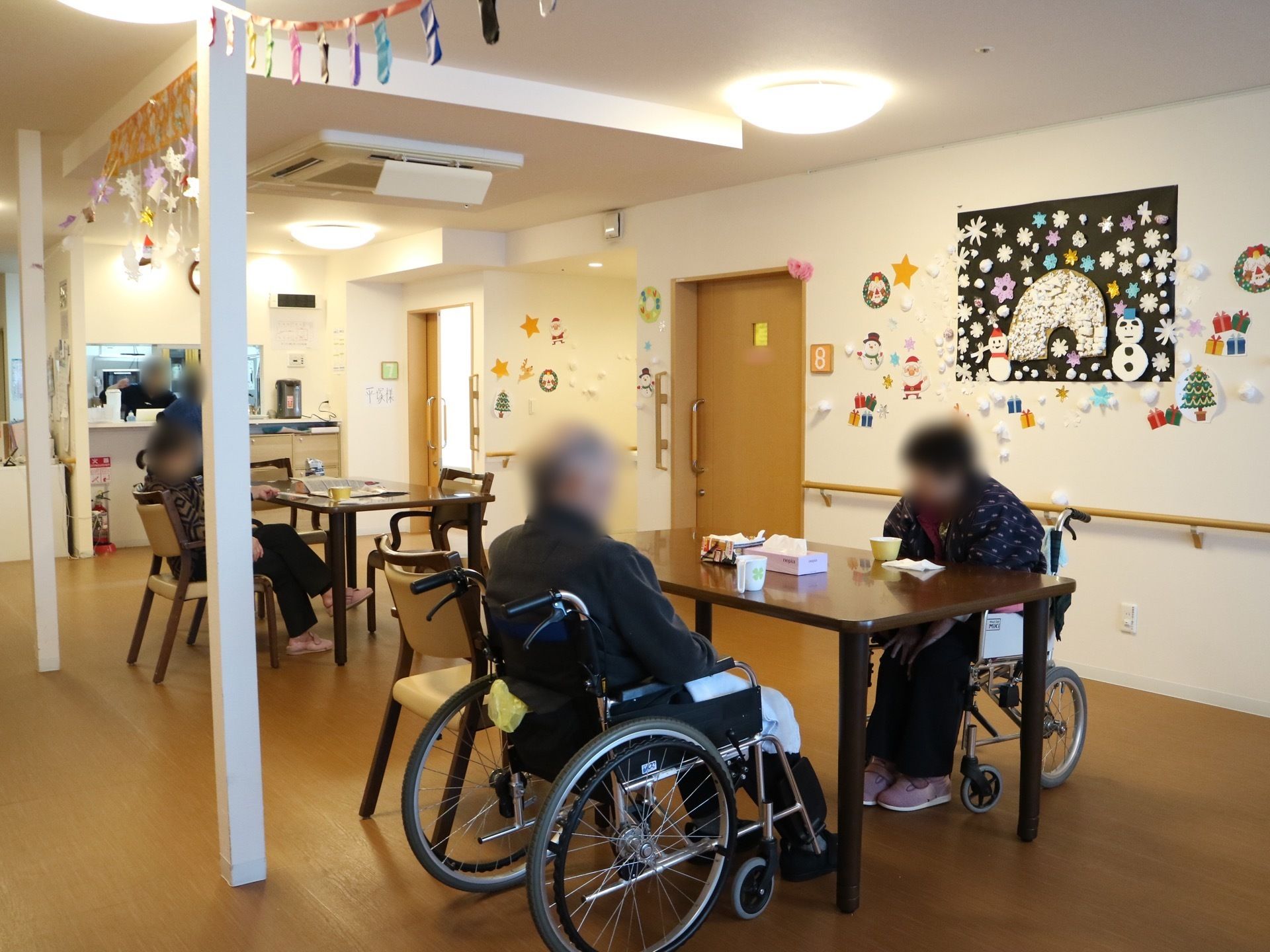 食堂のテーブルに、車椅子をご利用の方が、車椅子に腰かけたままで向かい合って座っている。白い柱が２本立っている。