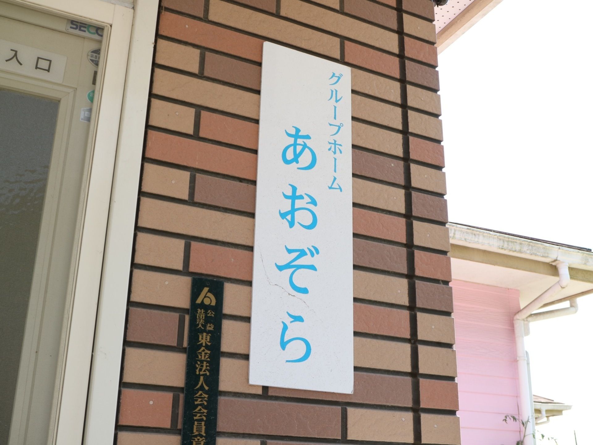 玄関前の外壁はレンガ張りで、白色に青い文字で書かれた表札と黒い表札の２枚が掛けられている。扉には、ぼかしの入ったガラスが使用されている。