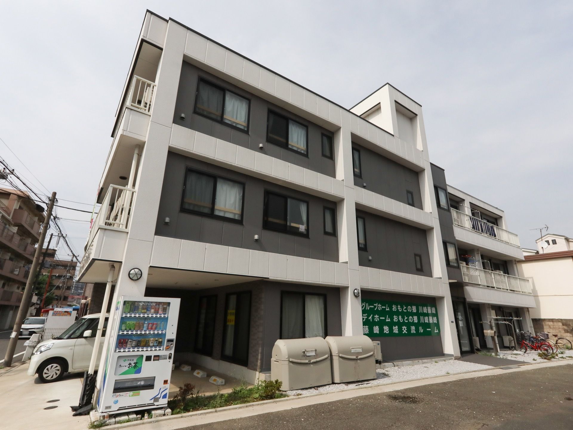 建物の１階に駐車スペースを完備している３階建ての施設である。１台の乗用車が停車しており、そばに自動販売機が設置されている。
