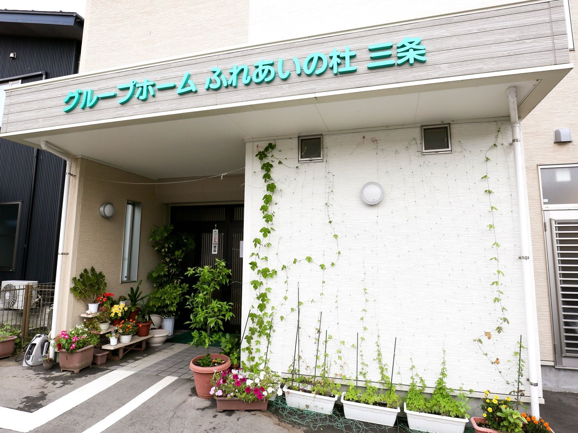 施設入口がある１階には小さな屋根があり、緑色で施設名が表示されている。屋根の下の壁には、つる性植物の葉が広がっている。