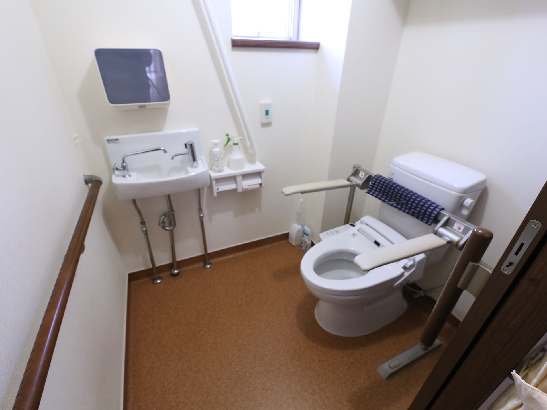 便座の両側に折り畳み式の手すりが取りつけられたトイレが用意されている。奥の壁際に洗面台とトイレットペーパーホルダーがある。