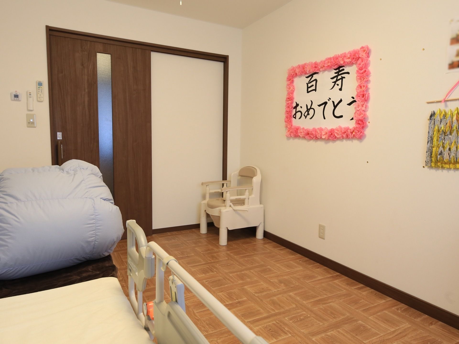 居室の入口はダークブラウンの木製の引き戸になっている。入口前に白い椅子が置かれ、壁に百寿のお祝いの飾りがされている。