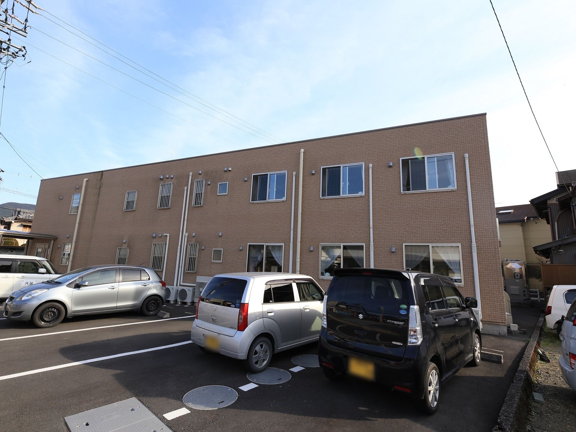 茶色の外壁の施設で、２階建てである。居室の窓が並び、排水管が通っている。施設前に駐車スペースが広がっている。