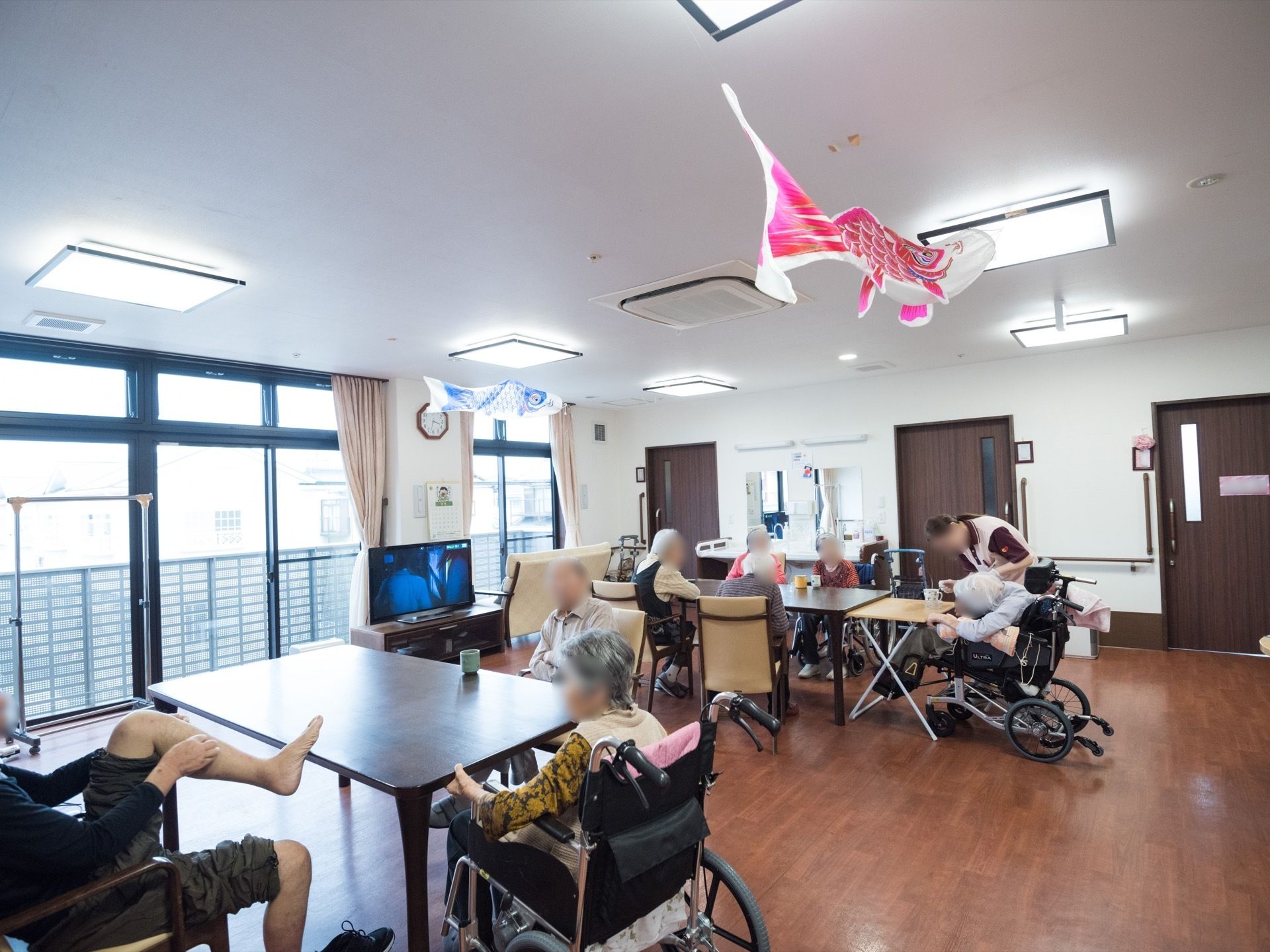 入居者様たちがリビングに集まりテーブルを囲んで、思い思いの時間を過ごしている。窓際にはテレビが１台設置されている。