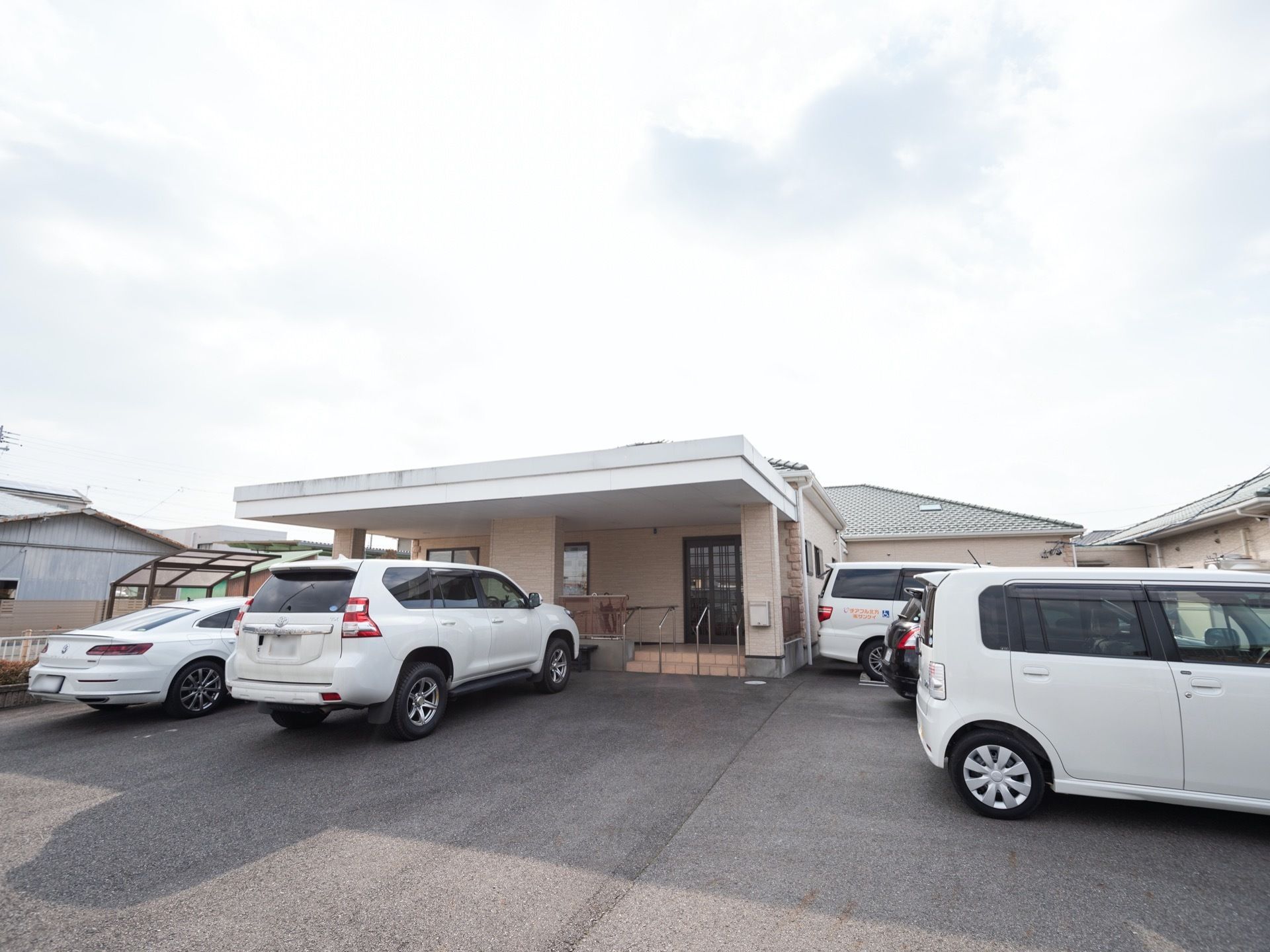 平屋建て施設の周囲の駐車スペースには、乗用車が複数台停車している。テラス屋根が装備されたスペースが設けられている。