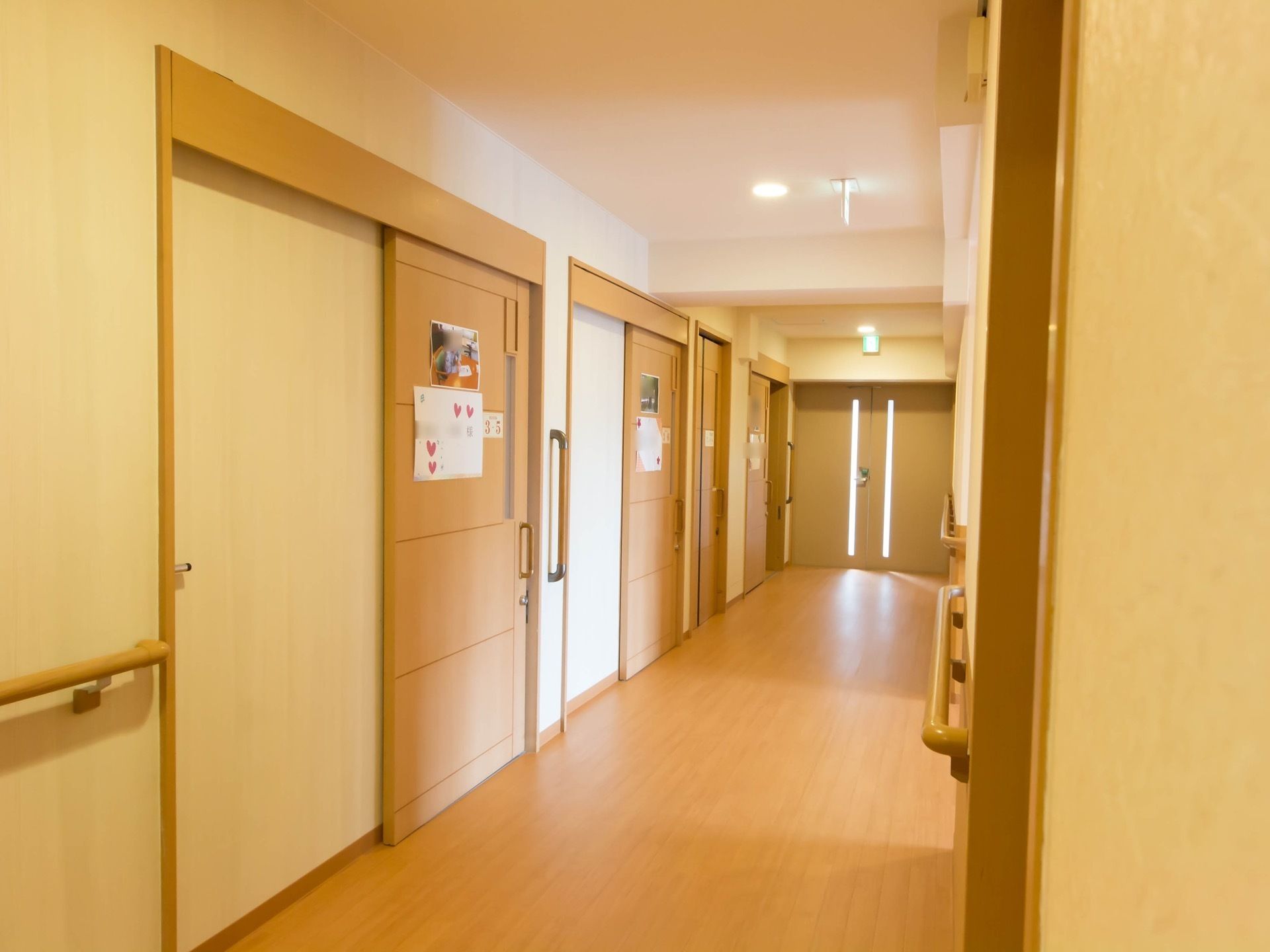 白い壁に床と同系色のベージュ色の居室扉が並んでいる。扉は大きく吊下げタイプの引き戸である。扉の隣の壁には縦向きの手すりがある。