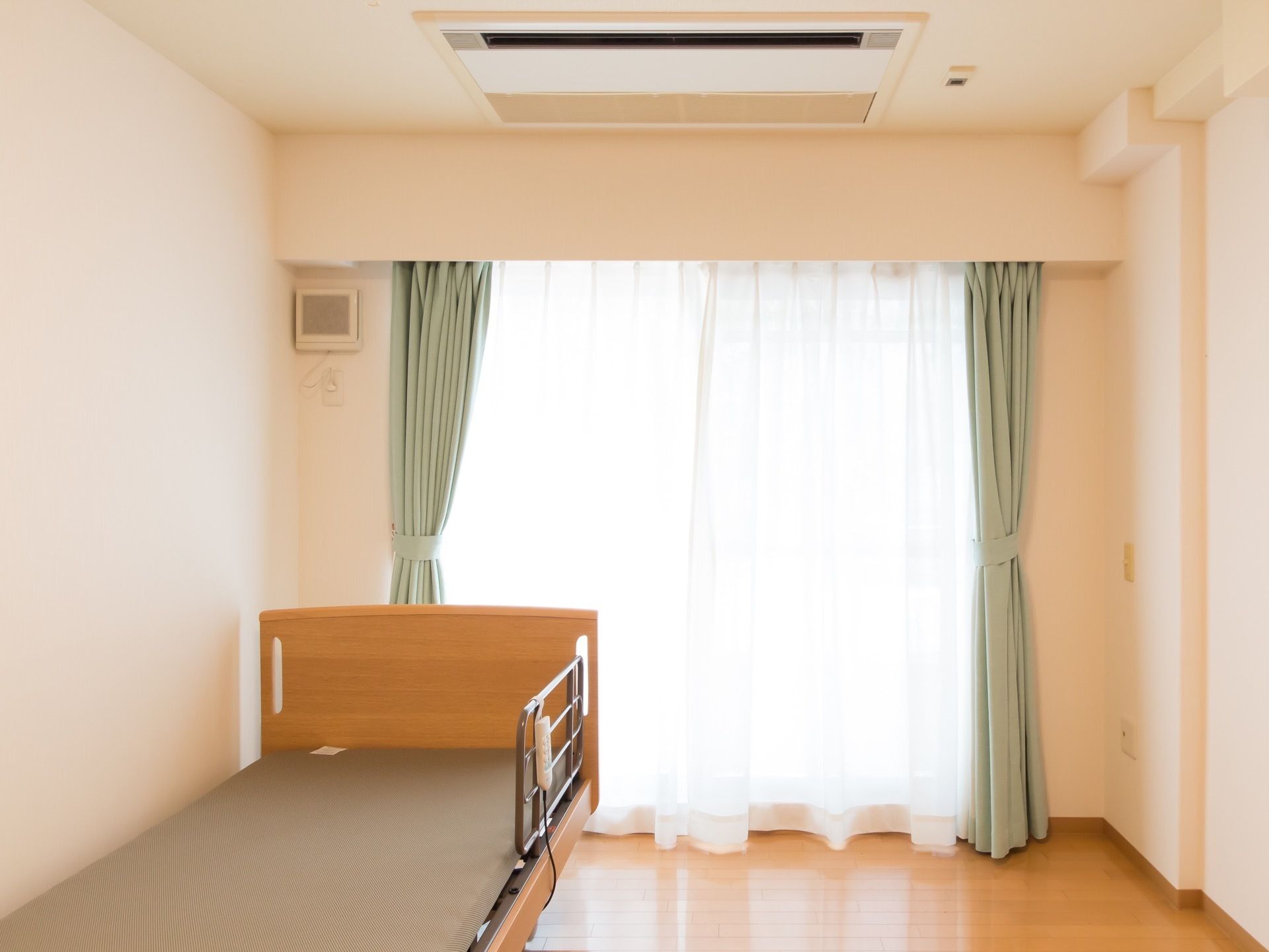 居室の正面は、壁面いっぱいにガラス戸があり日が射し込んでいる。天井には、空調を管理する機器が設置され、壁面際にベッドを用意。