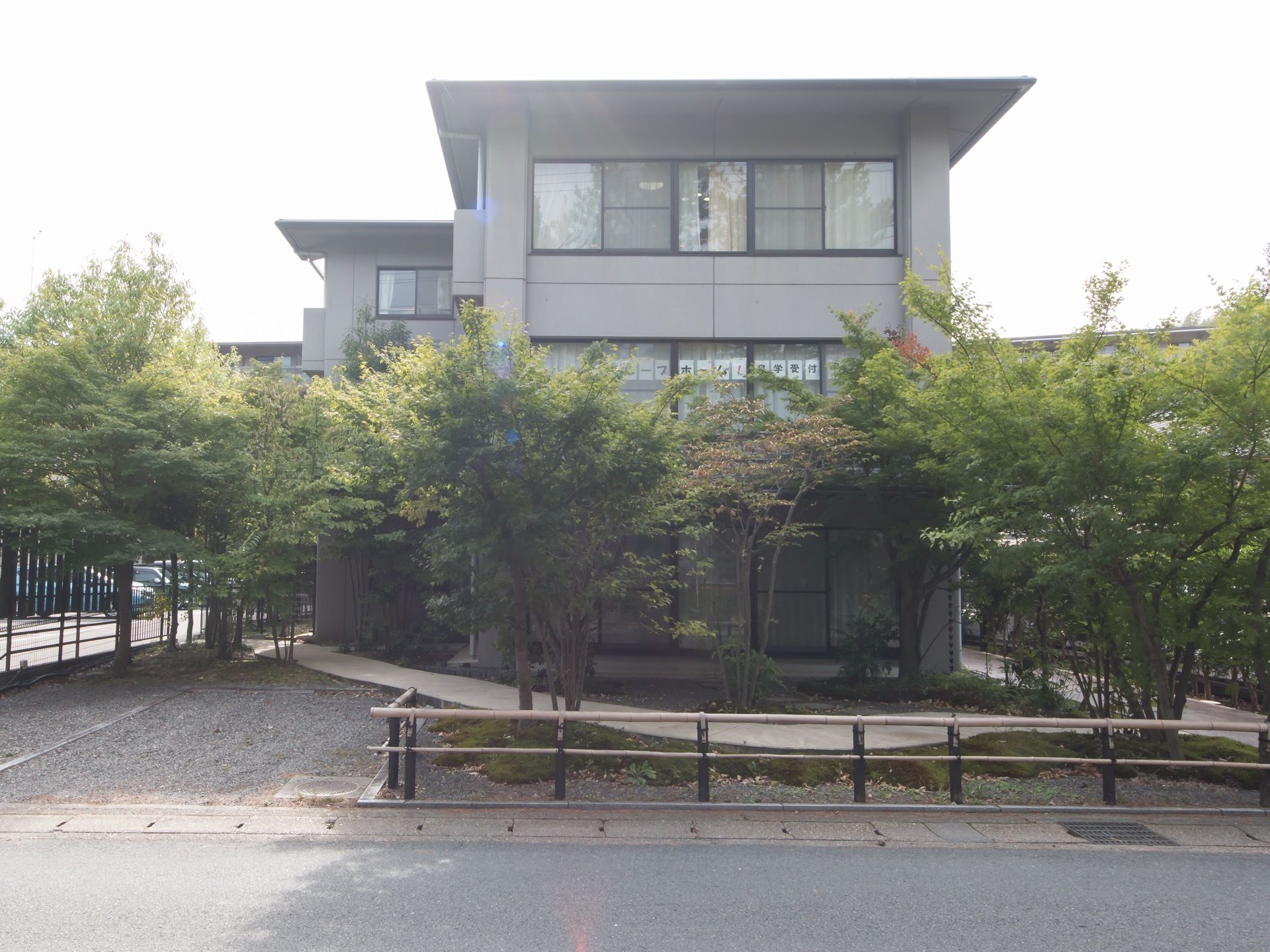 グレーの外壁の３階建ての施設建物である。窓には張り紙がしてあり、建物は道路に囲まれた場所に建っている。