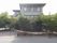グレーの外壁の３階建ての施設建物である。窓には張り紙がしてあり、建物は道路に囲まれた場所に建っている。