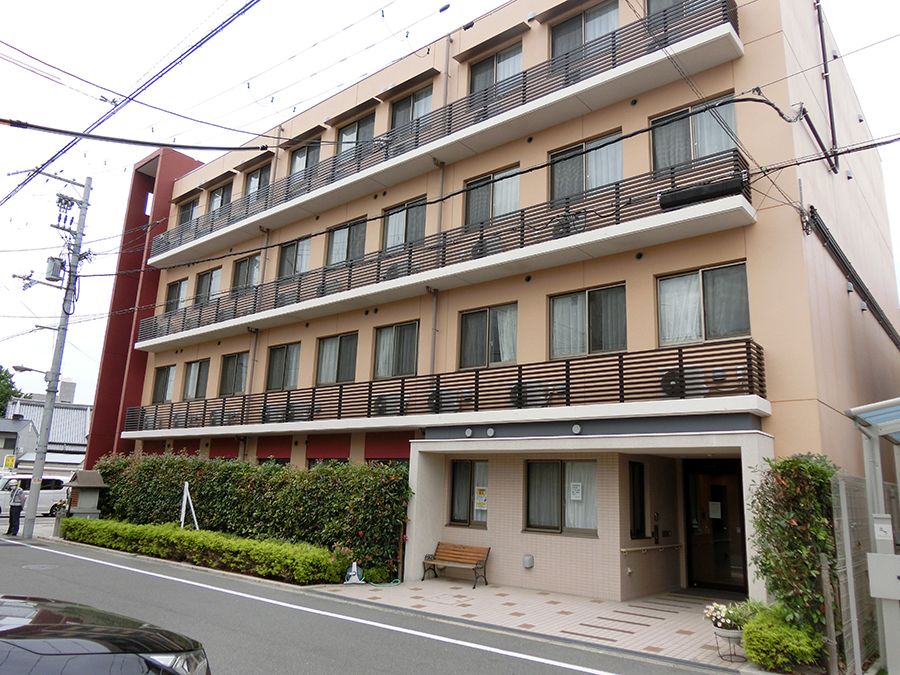 緑が周囲に植えられたブラウン系の外壁の４階建ての建物。玄関にはベンチも置かれている。