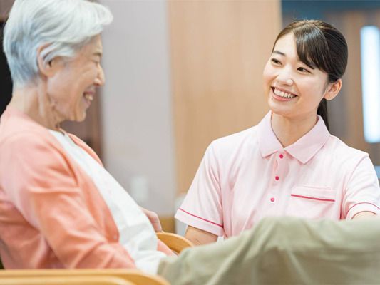 ピンクのポロシャツを着た若い女性スタッフと白髪の高齢女性が笑顔で話している。