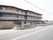 ３階建ての建物で、壁は木目調になっている。建物の前には駐車場があり、車やバイクが停められ、白いフェンスが建てられている。