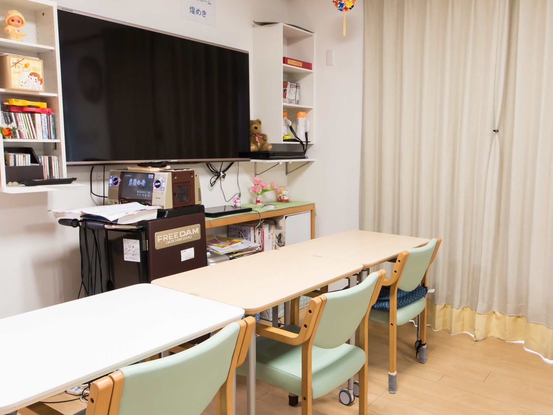大型のテレビが壁に取り付けられ、その手前にテーブルとひじ掛け付きの椅子が複数設置されているのが見える。