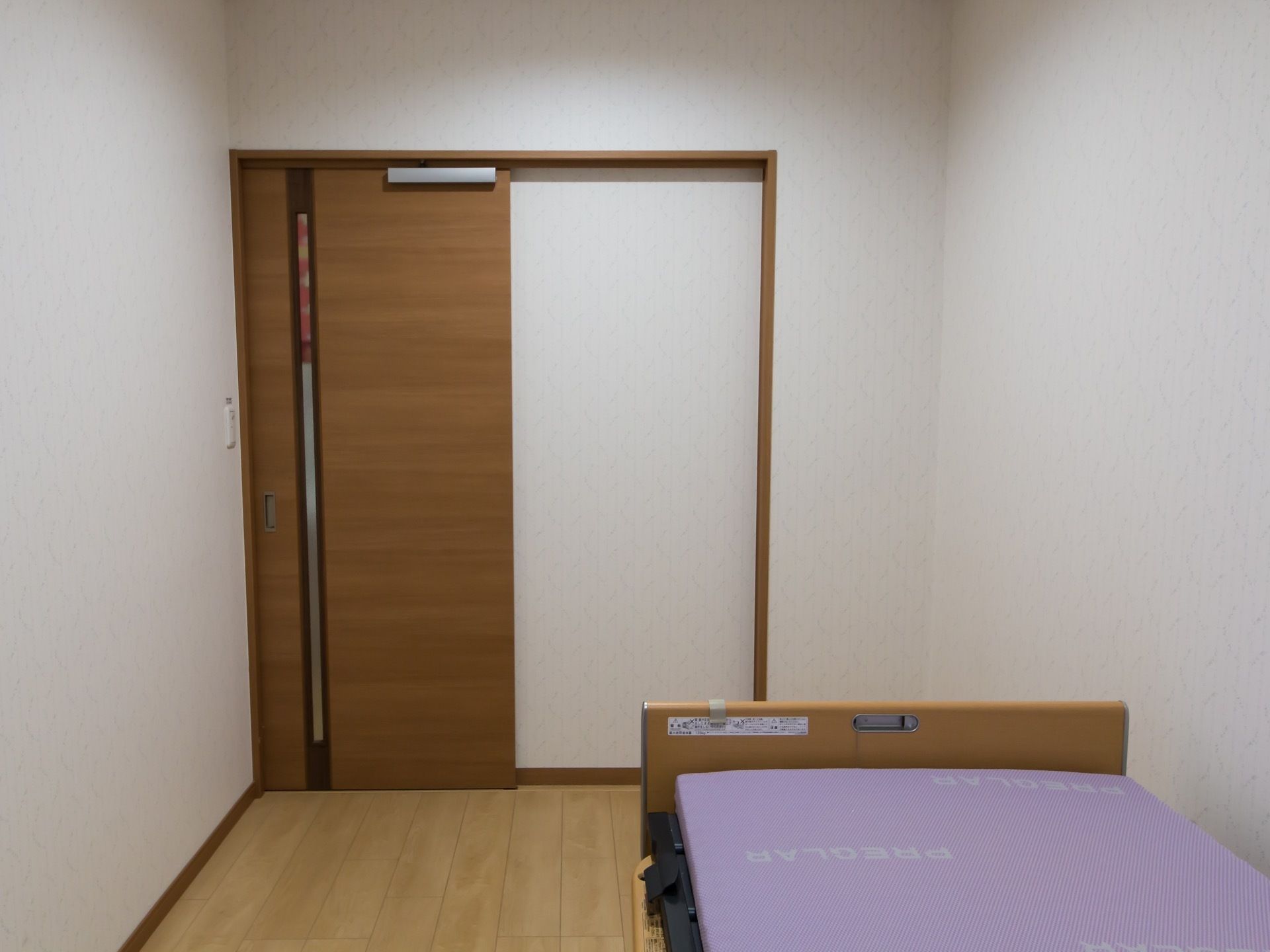 居室の壁は白色で、床はフローリングである。部屋の扉は木製でスライド式の物が取り付けられている。1台のベッドが置かれている。