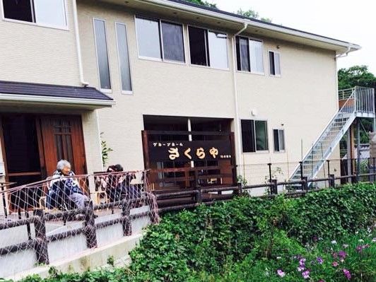屋根が付いた玄関前の椅子には入居者が2名座って話をしている。2階建ての建物はベージュ。