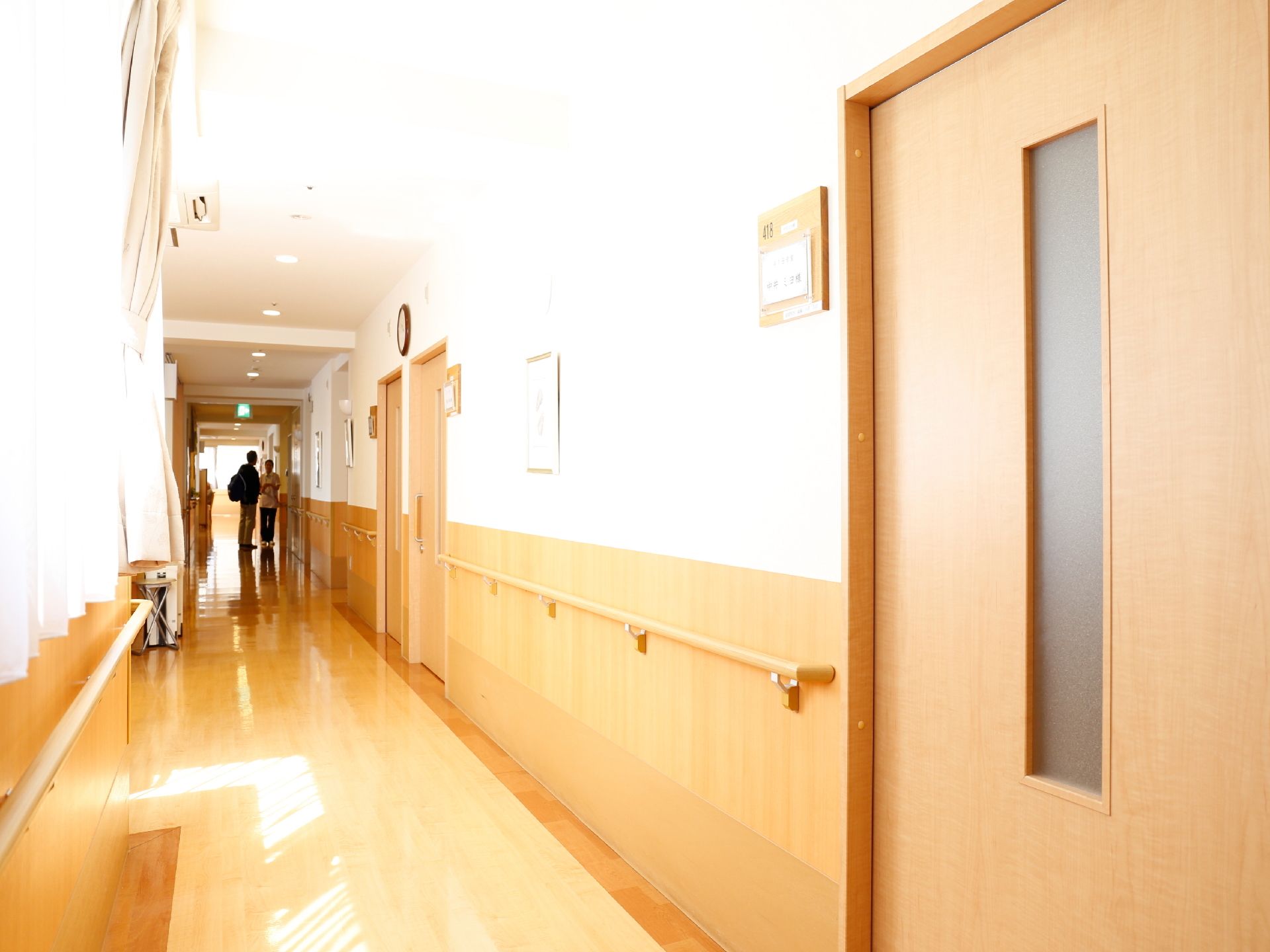 施設内の廊下は、フローリングの床と白い壁の温かみのある内装になっている。右側には、引戸の取りつけられた居室が並んでいる。