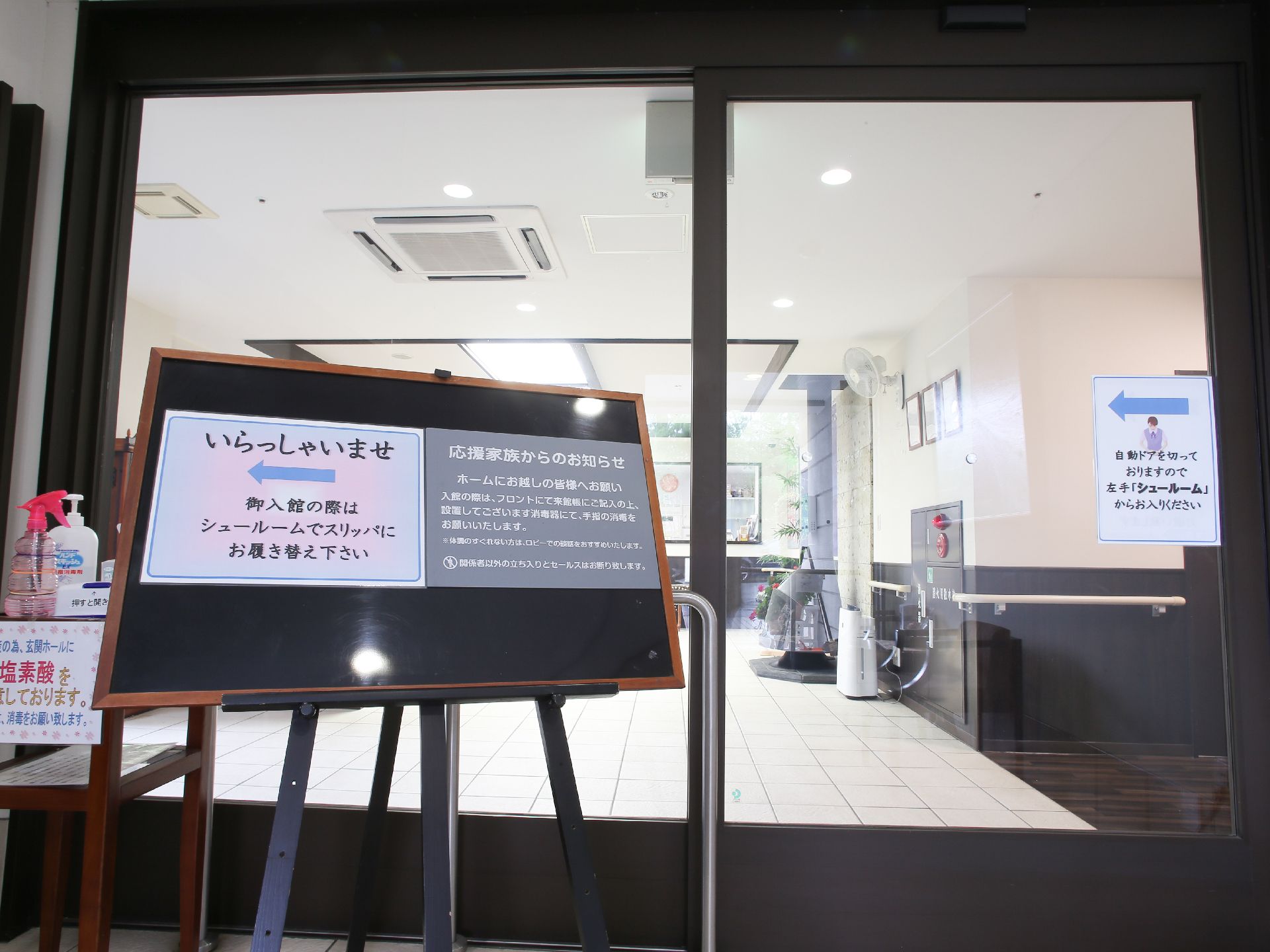 入り口に大きな自動ドアがある。下駄箱やフロントへの案内看板が置いてある。手の消毒薬も置いて館内への感染対策をしている。