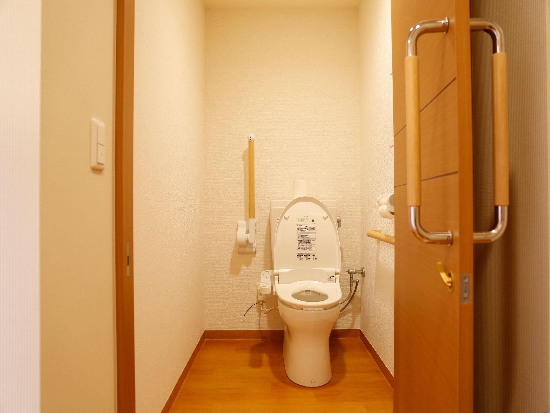 居住者の個室に設置されたトイレ。木製の扉を開けると洋式便器が見え、利用者を補助する手すりが壁に固定されている。