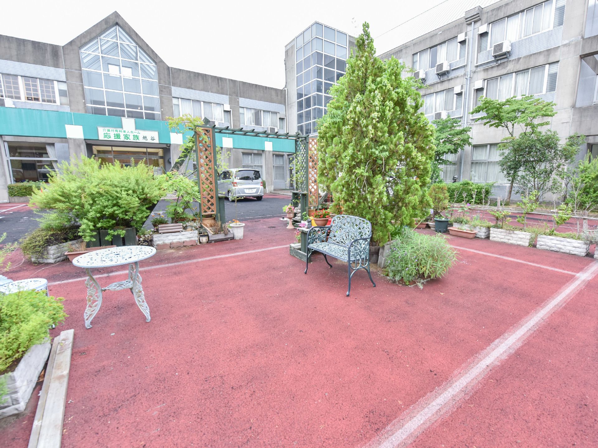 テラスの向こうに駐車場と施設が見える。緑がいくつも植えられており施設は外観にガラス戸が多く使われている。