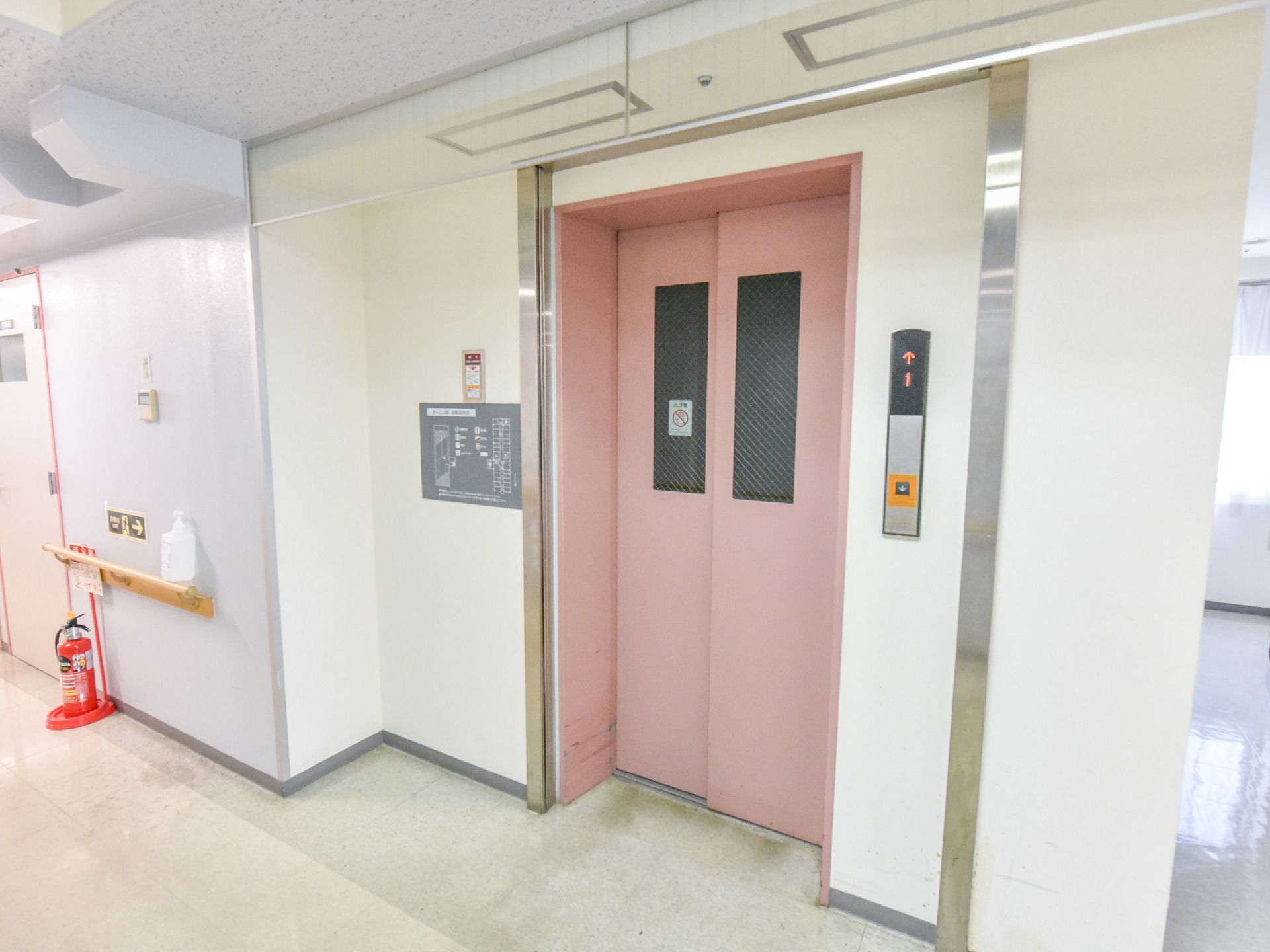 壁には手すりが設置され下には消化器が置かれている。ピンク色のエレベーターの扉は2枚構造になっている。