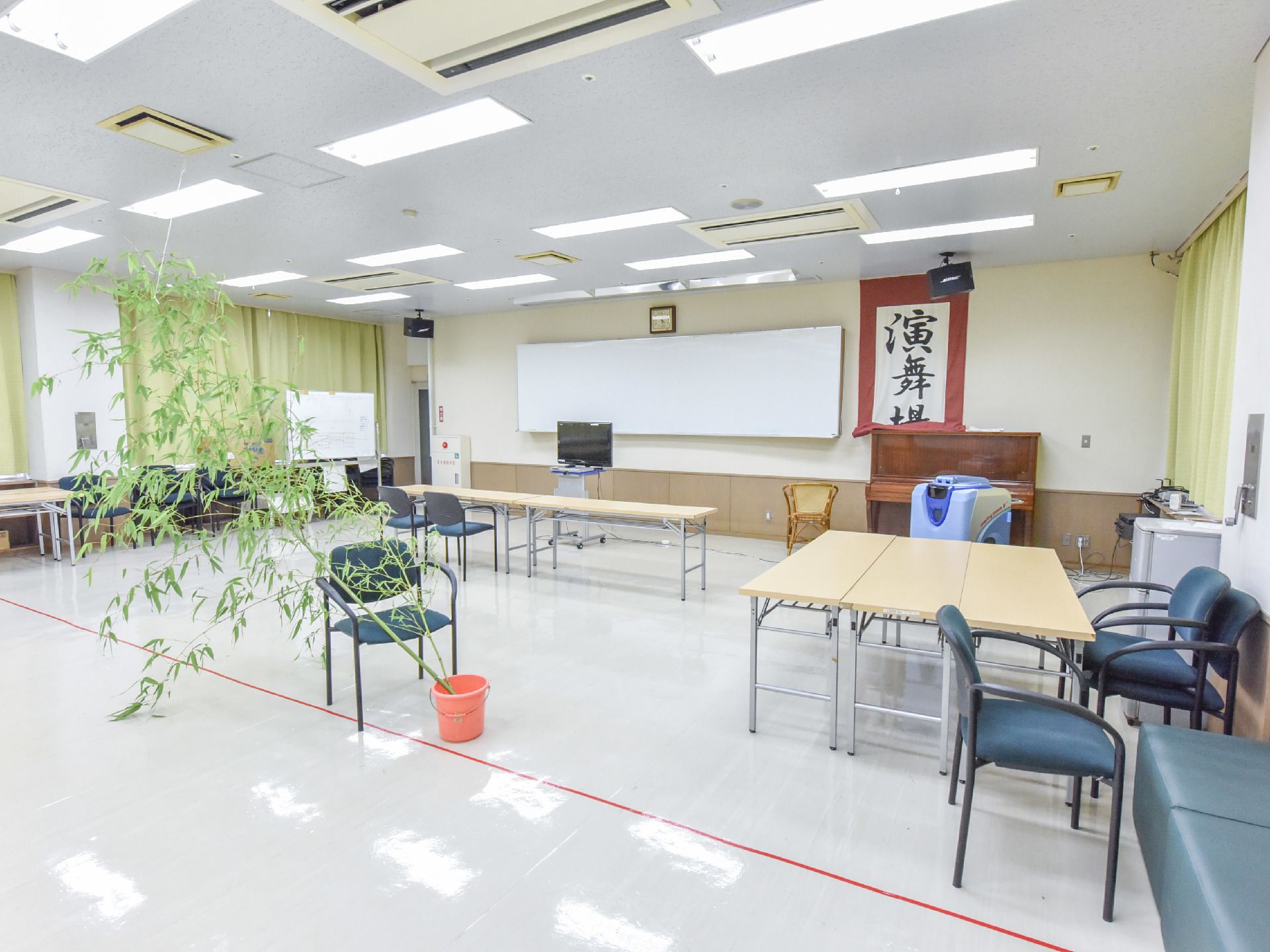 奥の壁には掛け軸やホワイトボードがかかっている。長テーブルがいくつも置かれており椅子やソファなども壁際に寄せられている。