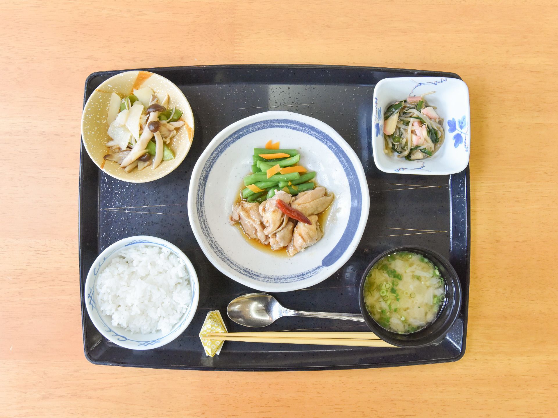 肉と野菜の主菜を中心に、キノコなどのおひたしの小鉢が2品とご飯に味噌汁などの健康的な食事が並んでいる。