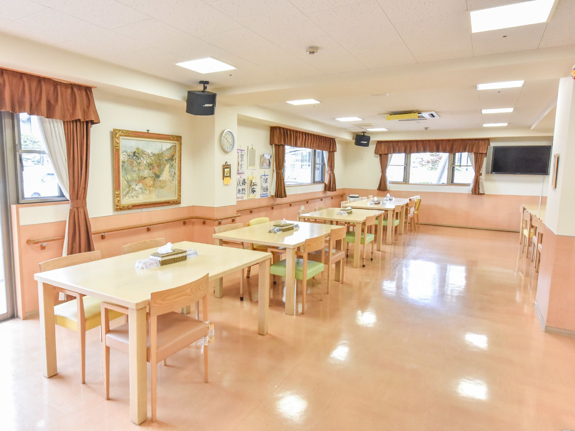 四角い木製のダイニングテーブルが5つある。シート部が緑色の椅子が20数脚ほどある。腰高窓、出窓がある。絵画がある。テレビがある。