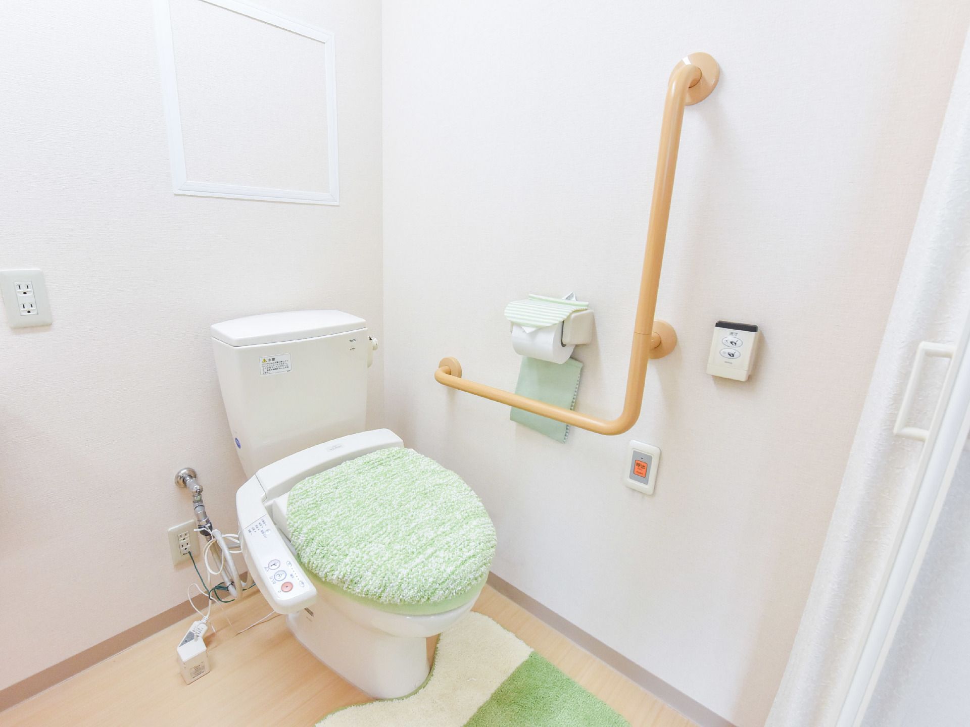 トイレの扉はアコーディオンカーテンである。床に緑色のマットが敷いてある。洋式トイレがある。壁に水を流すためのリモコンスイッチがついている。
