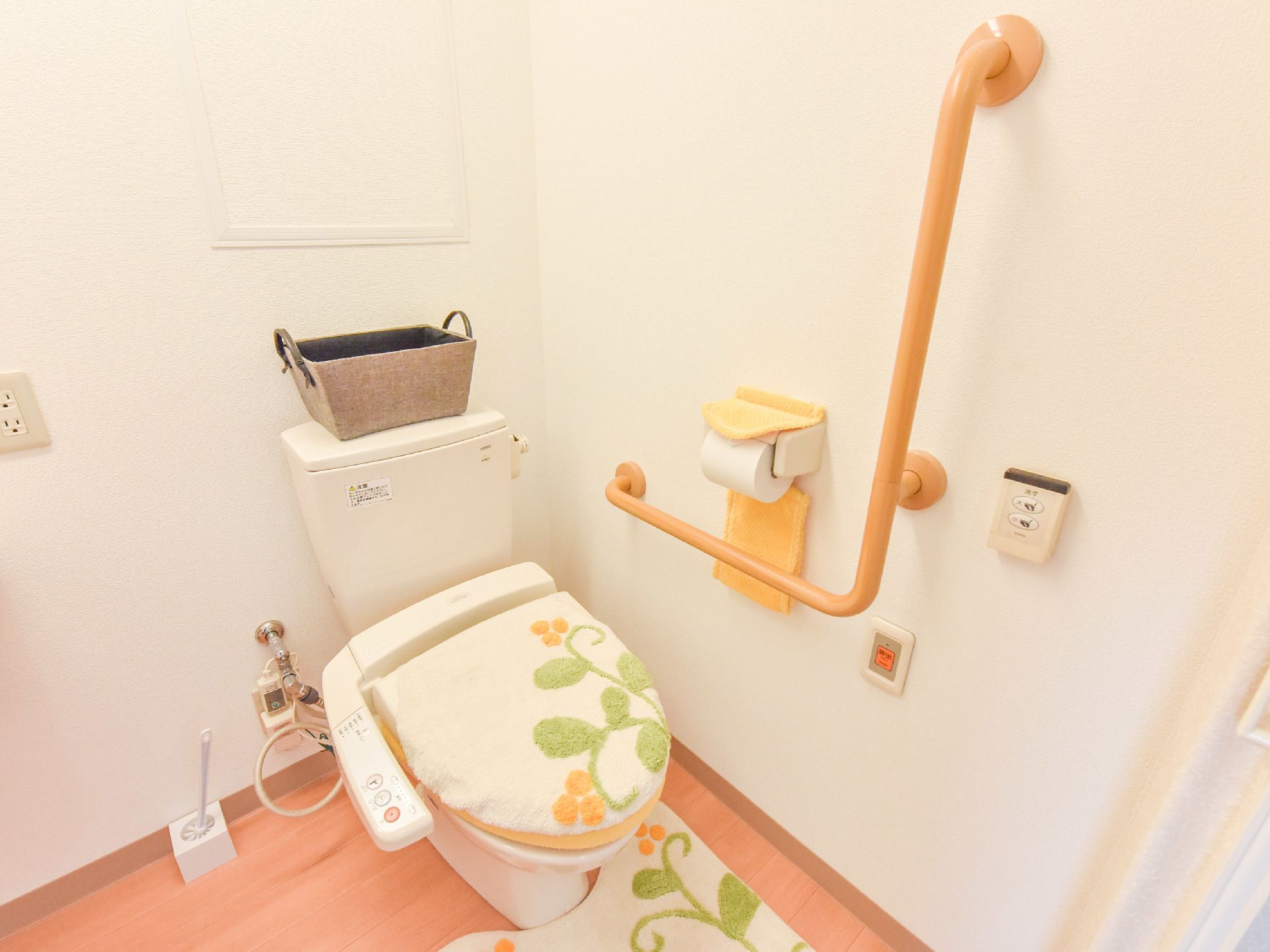 茶色の床に、白い壁のトイレである。トイレの掃除用ブラシがある。ウォシュレットタイプの洋式トイレである。手すりそばに、呼び出しブザーがある。
