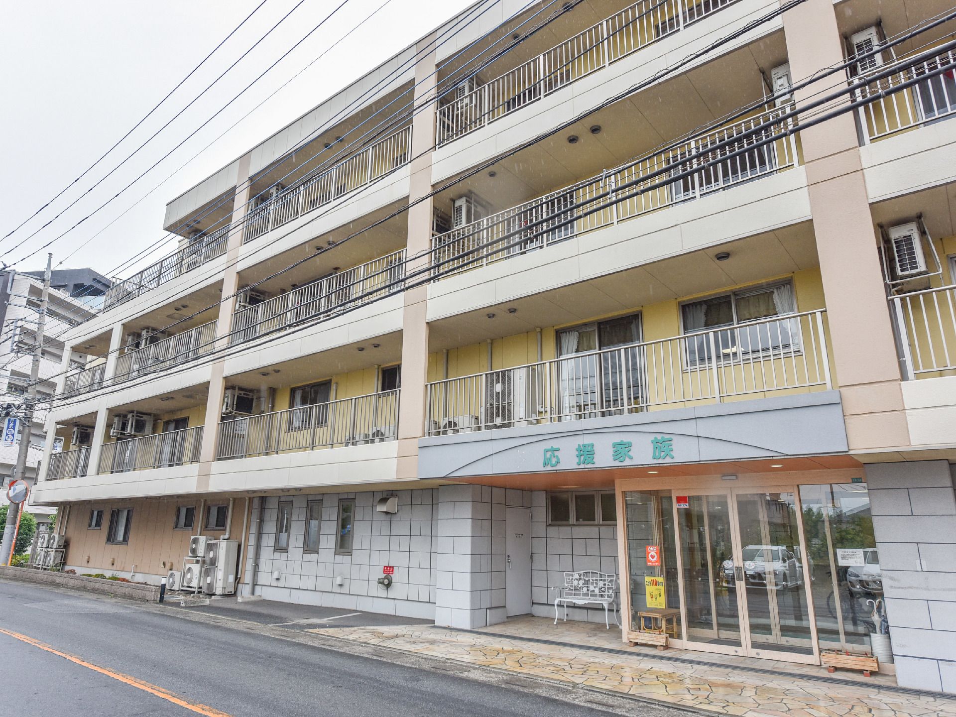 1階部分はグレイと茶色、2階から4階にかけては黄色い色の壁である。花壇があり低木が植えてある。玄関前に白い鉄のベンチがある。