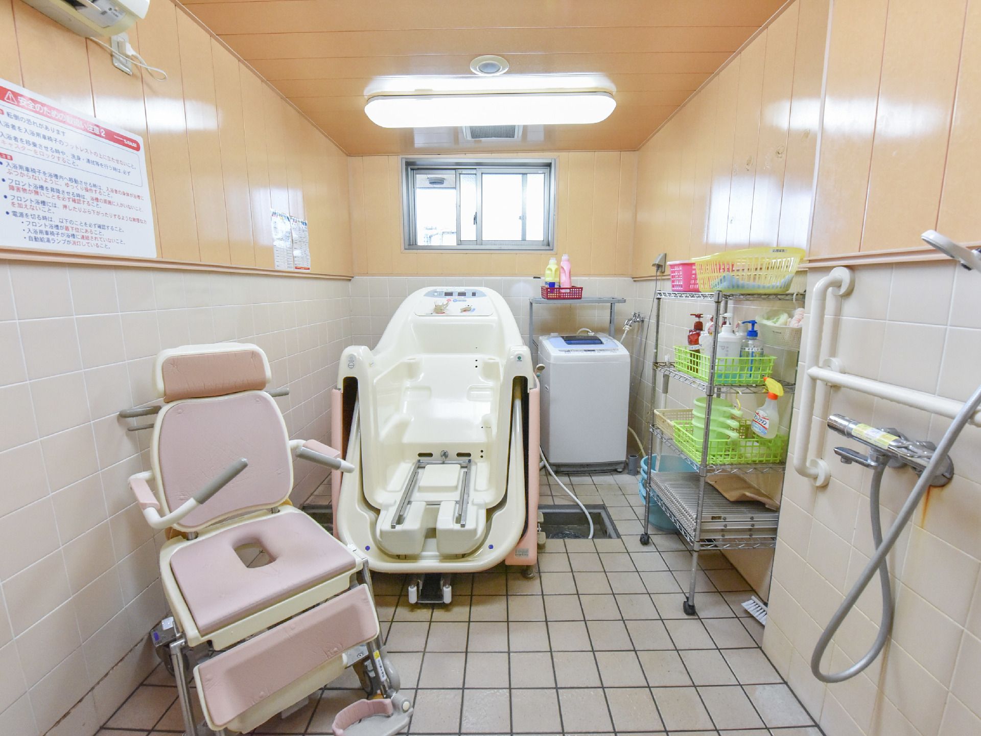 窓のあるタイル張りの部屋に入浴用車いす、バスタブ、洗濯機、シルバー色の棚が置いてある。棚には、籠がたくさん置いてあり、湯桶がある。