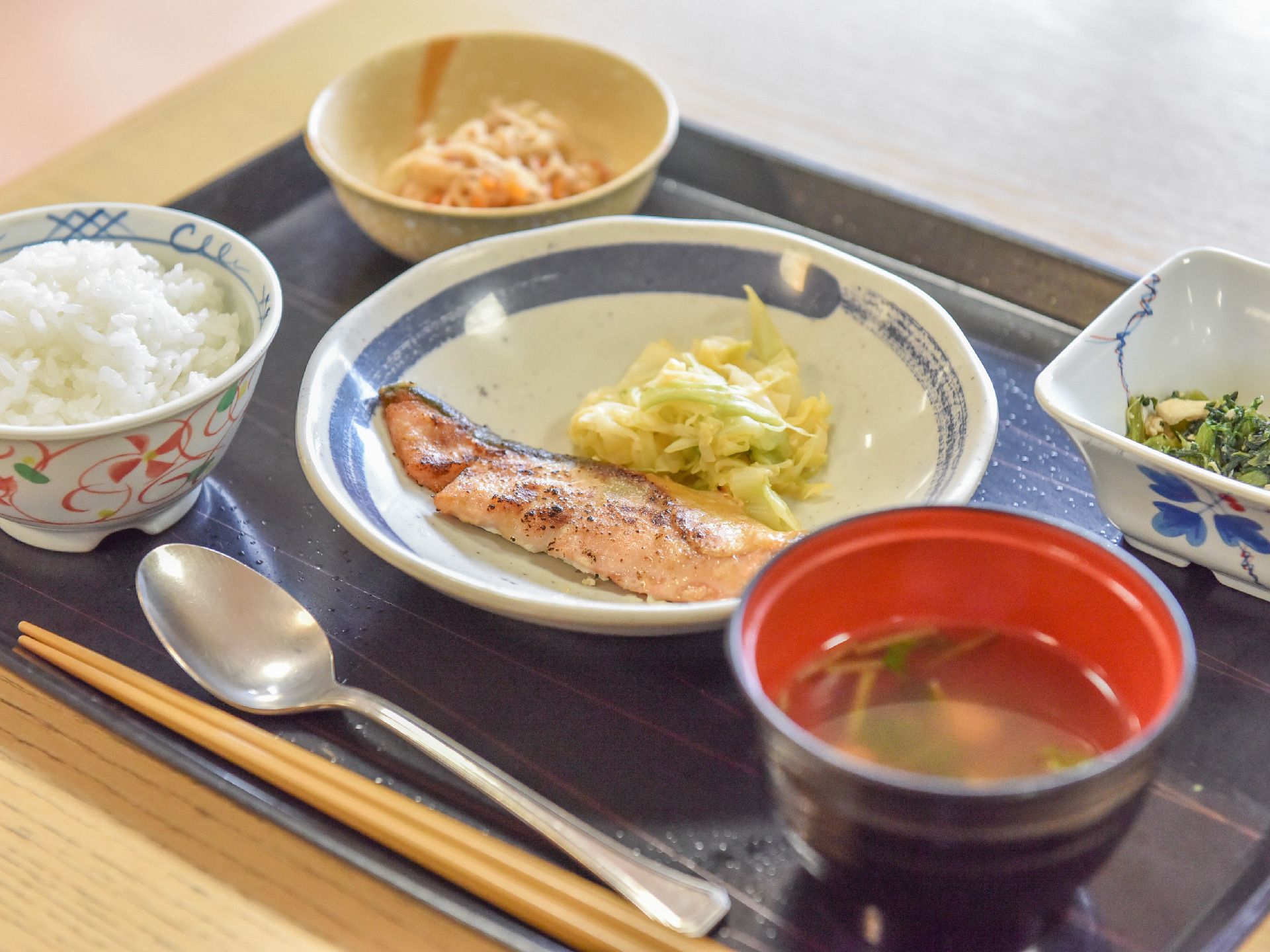 木製テーブルの上に黒いお盆がある。箸、スプーンが盆に載っている。ごはん、味噌汁、魚、小鉢が2つついていて、合計5点の食事である。