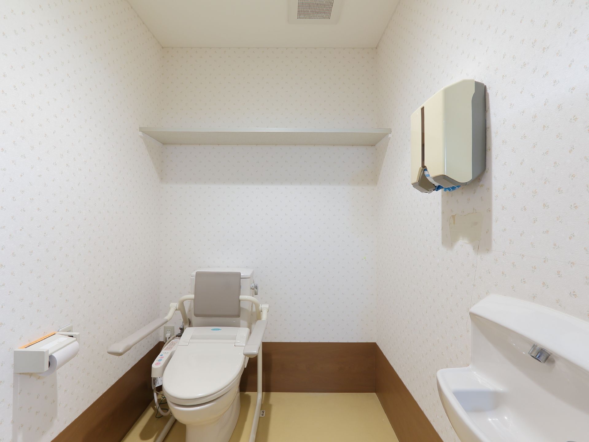 柄がついているが、白っぽい壁紙のトイレで、床はクリーム色である。洋式トイレは白で、貯水タンクに背あてクッション、左右に手すりがついている。