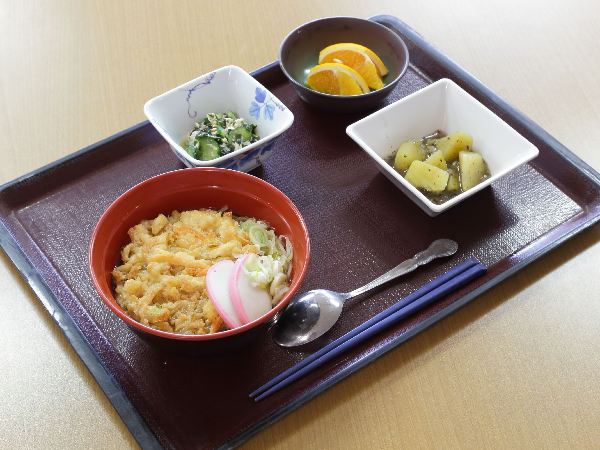 食事の一例である。盆の上に四角い深鉢が2つ置いてある。丸い深鉢にはオレンジが入っている。ごはんは天丼である。スプーン、箸の両方が置いてある。