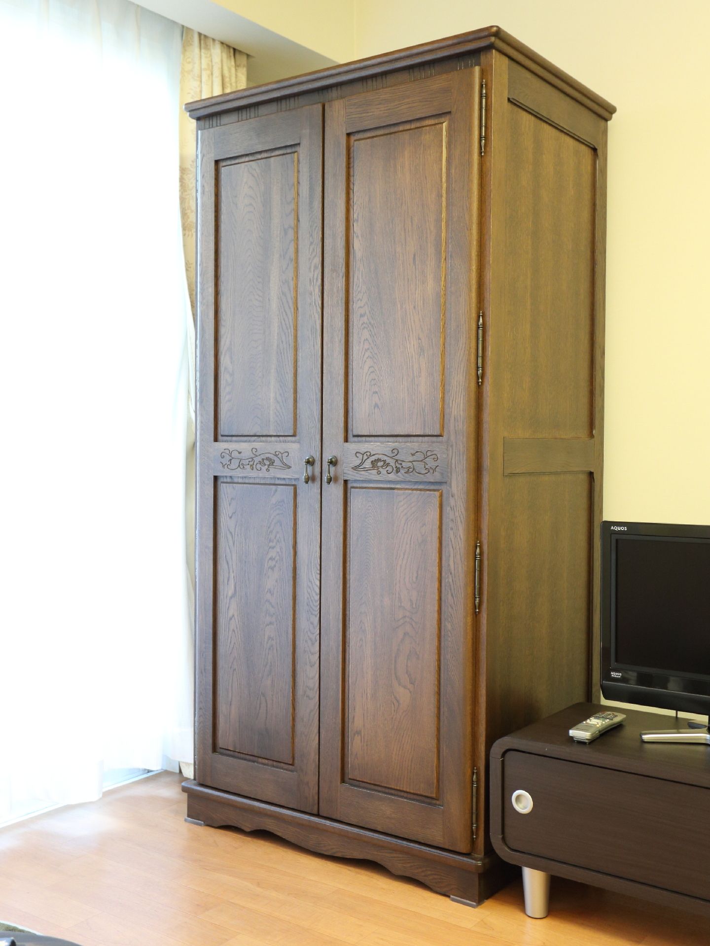 フローリングの部屋である。茶色のクローゼットがある。隣にテレビとテレビラックが置いてある。窓にレースとカーテンが下げてある。
