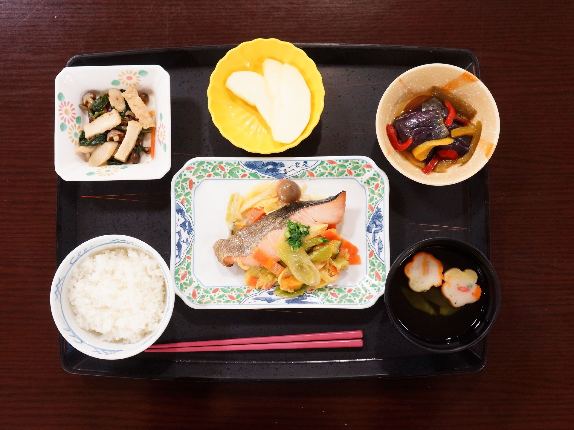トレイの上に載っている食事の一例である。赤い箸一膳。ごはん、吸い物のほか、メインの惣菜、副惣菜、フルーツがある。